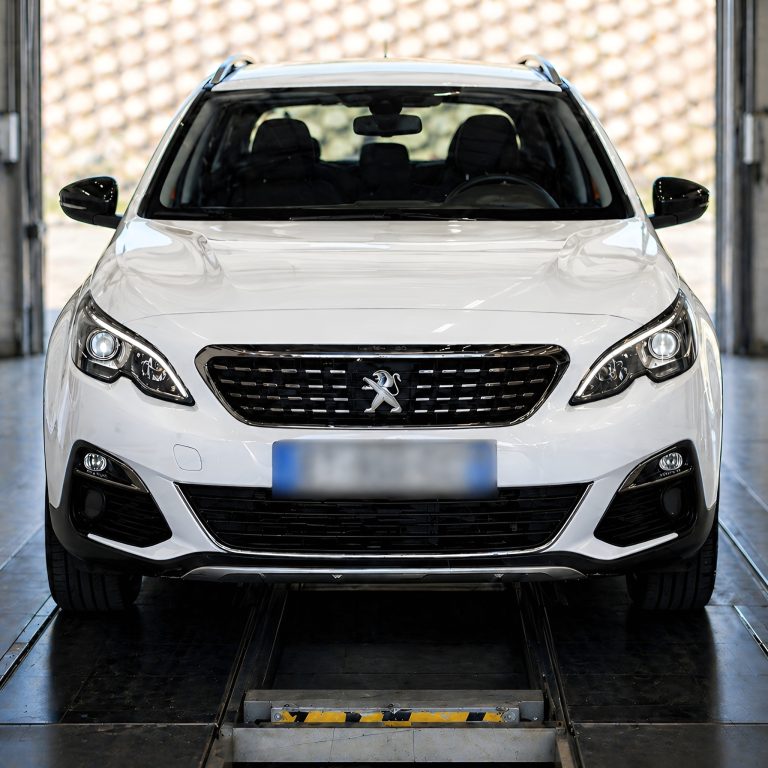 Peugeot blanche photographiée dans un centre de contrôle technique automobile, positionnée sur la zone d’inspection dans un garage moderne, avec plaque d’immatriculation floutée.
